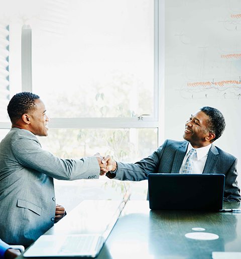 Two men shaking hands at a table in front of a laptop.